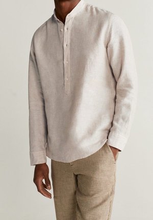 Mężczyzna w beżowej lnianej koszuli z długim rękawem i zapinanym na guziki przodzie oraz w brązowych spodniach w kratę, jedna ręka w kieszeni.