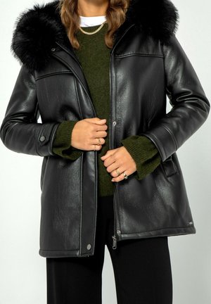 Femme portant une veste en cuir noire avec col en fourrure sur un pull vert, tenant les bords de la veste près de la fermeture éclair et des boutons.
