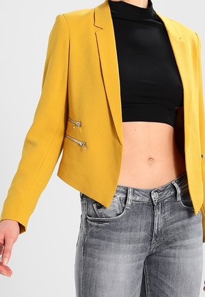 Vrouw draagt een mosterdgele cropped blazer met twee ritszakken, een zwart cropped top en vervaagde grijze spijkerbroek tegen een effen achtergrond.