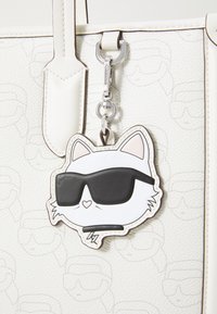 White tote bag with embossed cat outlines features a cat-shaped keychain in black and white, wearing black sunglasses and outlined in black.