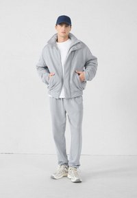 Lichtgrijze zip-up jas met een verhoogde kraag en drukknoopsluiting, gecombineerd met bijpassende joggers, een wit T-shirt en grijze sneakers.
