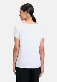 Biała bluzka z krótkim rękawem o szerokim dekolcie i gładkiej teksturze, charakteryzująca się prostym krojem i detalem w postaci zakładki z tyłu.