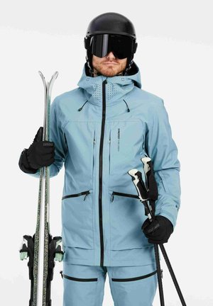 Hellblauer Skijacke mit schwarzen Reißverschlüssen, verstellbarer Kapuze und Belüftung. Das Model hält Skier und Stöcke und trägt schwarze Handschuhe und einen Helm.