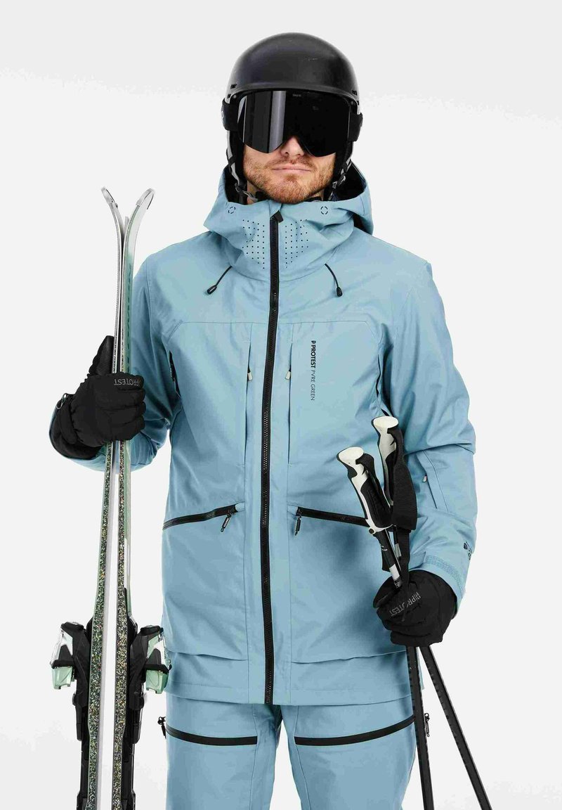 Lichtblauwe skijas met zwarte ritsen, verstelbare capuchon en ventilatie. Model houdt ski's en stokken vast, draagt zwarte handschoenen en een helm.