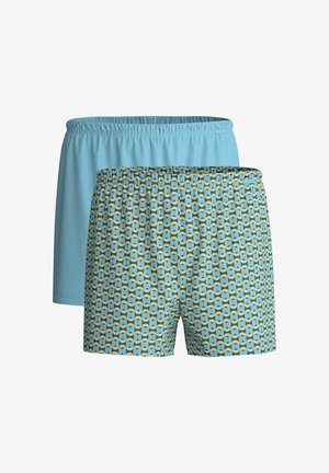Gespierde man met een lichte huid en krullend lichtbruin haar die een blauw, geel en zwart zwembroek met patroon draagt en staat tegen een simpele witte muur.