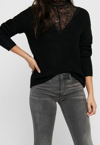 Pull noir en tricot avec un panneau en dentelle au niveau du col, associé à un jean gris slim. La texture est douce avec une coupe décontractée.