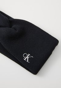 Gebreide donkerblauwe haarband met een textuurafwerking, voorzien van een voorste twist en een klein wit geborduurd logo aan de zijkant.