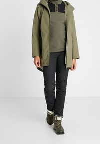 Veste imperméable vert olive avec capuche, intérieur en polaire noire, associée à un pantalon noir et des bottes montantes vert foncé à lacets avec doublure blanche.