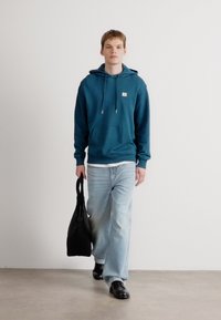 Tamsiai mėlyna palaidinė su reguliuojamomis raištelėmis, priekinė kišenė, baltas marškinėliai po ja, šviesiai mėlyni džinsai, juodi batai ir juoda vilkimo rankinė.