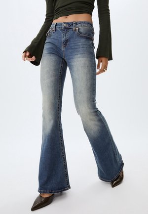 Kvinde iført falmede blå flare-jeans, mørkegrøn langærmet top og spidse sorte sko, stående foran en hvid baggrund.