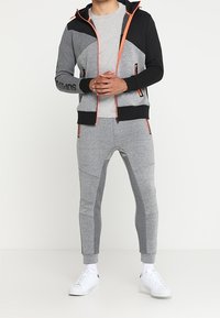 Grijze en zwarte trainingsoutfit met een rits hoodie, voorzien van oranje accenten en logo, gecombineerd met aansluitende grijze broeken en witte sneakers.