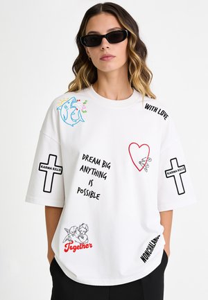 Donna che indossa una t-shirt bianca oversize con croci nere, squalo, cuore, angeli e frasi come "Dream Big Anything is Possible" e "Together."