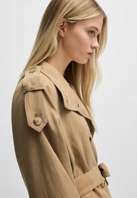 Beige trench coat with a wide collar, buttoned shoulders, and a belted waist. Made from a textured fabric with visible stitching details.