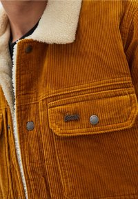 Giacca in corduroy arancione ruggine con colletto in sherpa crema. Presenta una tasca sul petto con un bottone a pressione in metallo e dettaglio del logo. Texture a righe verticali.