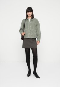 Giacca in corduroy verde con due tasche frontali, abbinata a una gonna a quadretti bianca e nera e collant neri. Tacchi neri completano l'outfit.