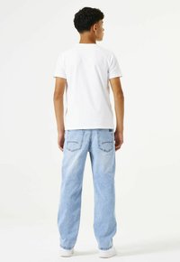 Camiseta blanca y vaqueros ligeros de tiro bajo vistos desde atrás, con costuras sutiles y dos bolsillos traseros con cremalleras. Zapatillas blancas.