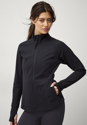 Kobieta w czarnej kurtce na zamek z otworami na kciuki, pozująca z jedną ręką w kieszeni na jednolitym jasnym tle.