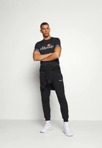 Černý sportovní outfit s tričkem s logem a tepláky. Obsahuje bílé detaily, elastický pas se šňůrkou a je doplněn bílými teniskami.