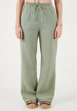 Nő széles szárú, világoszöld, zsinóros nadrágban és bézs szandálban, fehér háttér előtt állva.