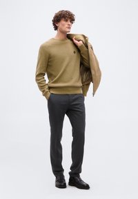 Άνδρας με σγουρά μαλλιά που φοράει πράσινο πουλόβερ και γκρι παντελόνι, κρατώντας ένα μπεζ σακάκι στον ώμο του, σε απλό φόντο.