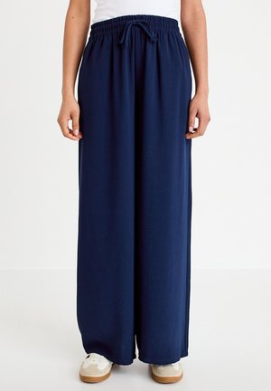 Osoba ubrana w luźne, granatowe spodnie o szerokich nogawkach z elastycznym pasem i białe trampki, stojąca na prostym, jasnym tle.