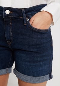 Tanssit denim-shortsit tummansinisinä, jossa on käännetty helma, etutasku, metallinapit kiinnityksessä ja kontrastiväriset ompelut. 