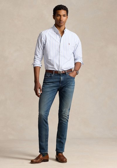 Man staat en draagt een wit-blauw gestreept overhemd met knopen, een blauwe spijkerbroek, een bruine riem, bruine loafers en een polshorloge, met één hand in de zak.