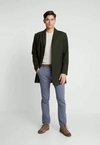 Homme portant un manteau vert foncé, une chemise blanche, un pantalon bleu, une ceinture marron et des chaussures marron, se tenant devant un fond uni.