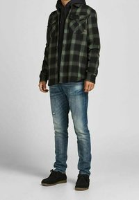 Camicia di flanella a quadri neri e verdi sovrapposta a un hoodie nero, abbinata a jeans chiari strappati e scarpe nere con suola in gomma.