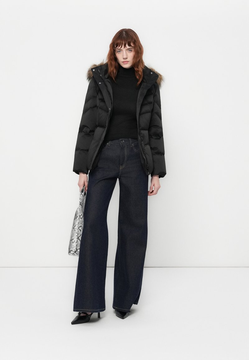 Mujer vestida con una chaqueta de plumas negra con capucha de piel, un cuello de tortuga negro, jeans oscuros de pierna ancha, tacones negros, mientras sostiene un bolso tote con estampado de serpiente.