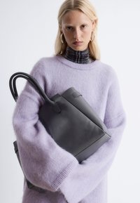 Black leather tote bag with curved handles, held against a light purple fluffy sweater, featuring a textured finish and clean lines.