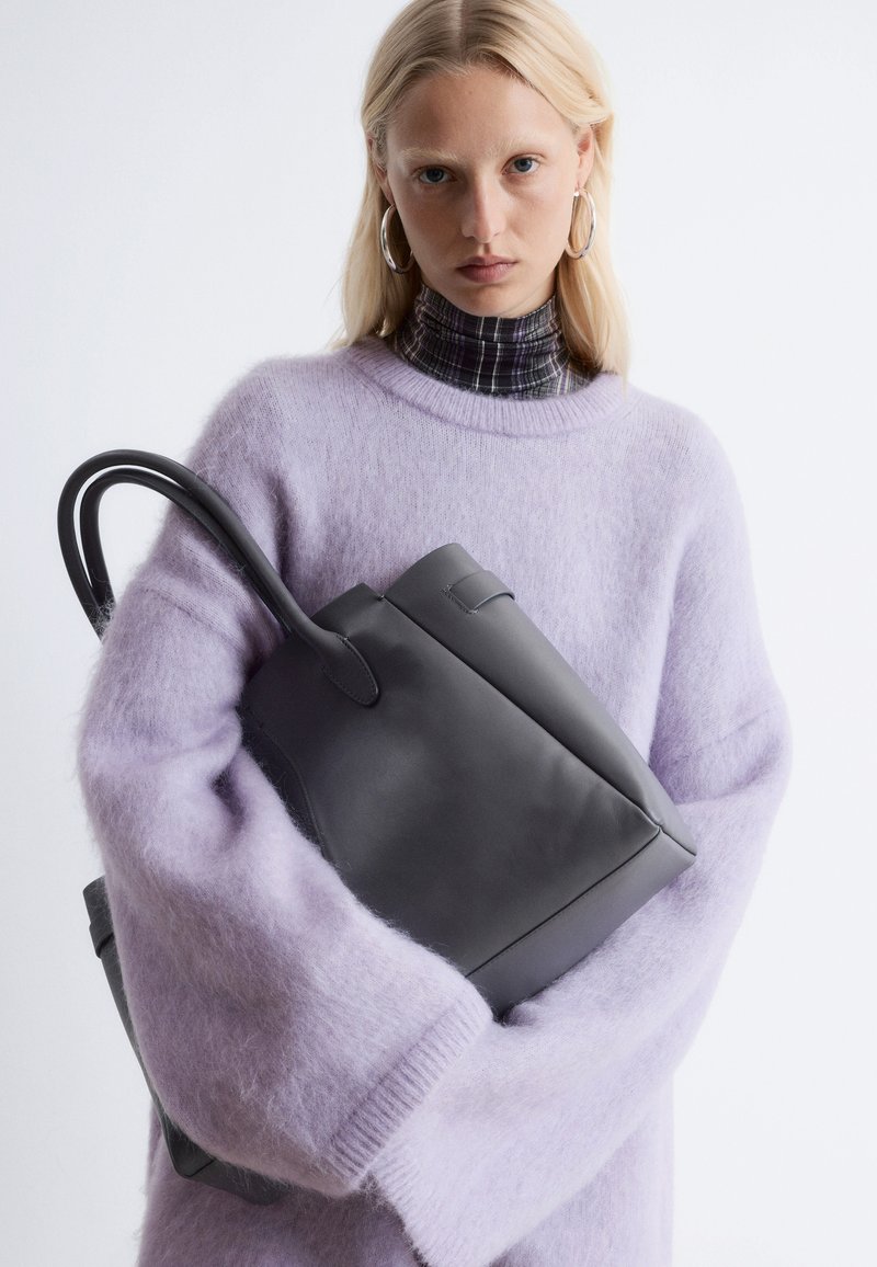 Black leather tote bag with curved handles, held against a light purple fluffy sweater, featuring a textured finish and clean lines.