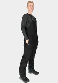 Black overalls with adjustable straps, zippered side pockets, and a sleek design. Paired with a dark long-sleeve shirt and black boots.