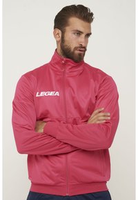 Röd idrottsjacka med hög krage, hel dragkedja och långa ärmar. Har en vit "LEGEA"-logotyp på vänster bröst och elastiska ärmar.