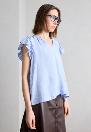 Vrouw met zwarte bril, een lichtblauw blouse met gerafelde mouwen en een bruine leren rok met knopen, staande tegen een effen muur.