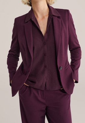 Kvinde iført en skræddersyet blommefarvet blazer, matchende knap-op skjorte og bukser med hænderne i lommerne, mod en neutral baggrund.
