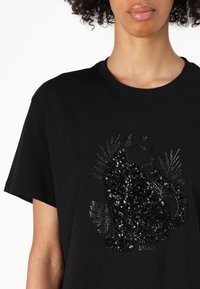 Maglietta nera in cotone con un design di un gatto realizzato con paillettes nere e dettagli argentati, su uno sfondo semplice.