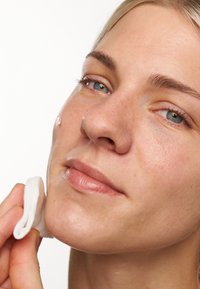 Nära bild av en person som använder en bomullsrondell på sin kind, som visar upp en mjuk hy, ljusreflektioner och minimal makeup.