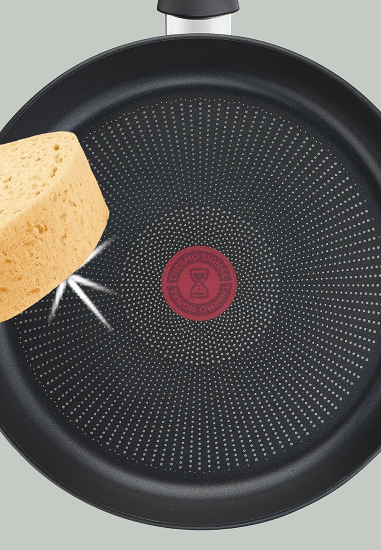 Padella antiaderente con superficie nera, indicatore rosso Thermo-Spot al centro e un pezzo di pane giallo sospeso sopra di essa.