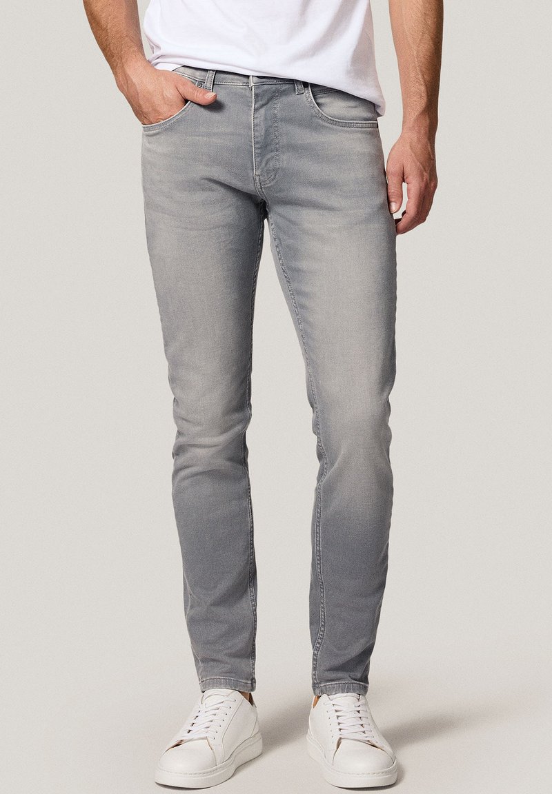 Mężczyzna w dopasowanych, wyblakłych szarych dżinsach z białymi trampkami i włożonej białej koszulce, stojący z jedną ręką w kieszeni.