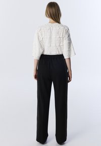 Top bianco a maniche corte con effetto stropicciato abbinato a pantaloni neri a gamba larga con decorazioni laterali e cintura elastica. Sfondo neutro.