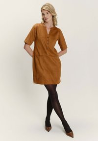 Krátkozrázový hnědý semišový šaty s knoflíkovou lištou a postranními kapsami. Model nosí černé punčochy a špičaté béžové podpatky. Hladká textura.
