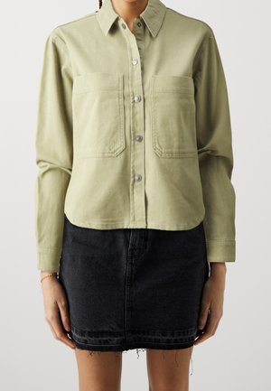 Femme portant une chemise boutonnée vert olive clair avec deux poches poitrine et une jupe en jean noire à ourlet effiloché, debout devant un fond uni.