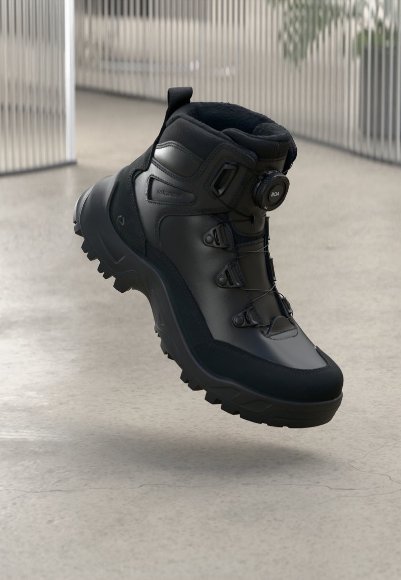 Bota de senderismo negra e impermeable con suela resistente y sistema de cordones BOA flotando sobre un suelo de concreto.
