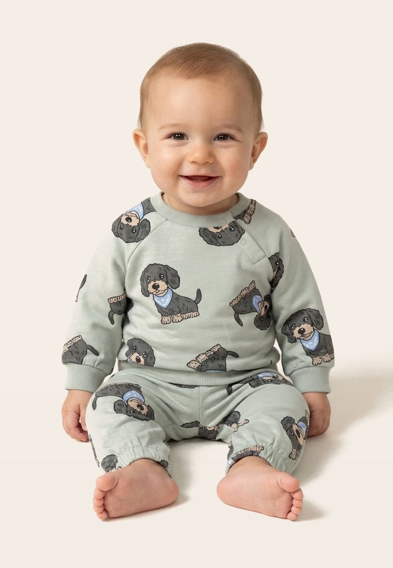 Bébé souriant assis, portant une tenue vert clair avec des imprimés de chiens noirs et des bandanas bleus, sur un fond beige uni.