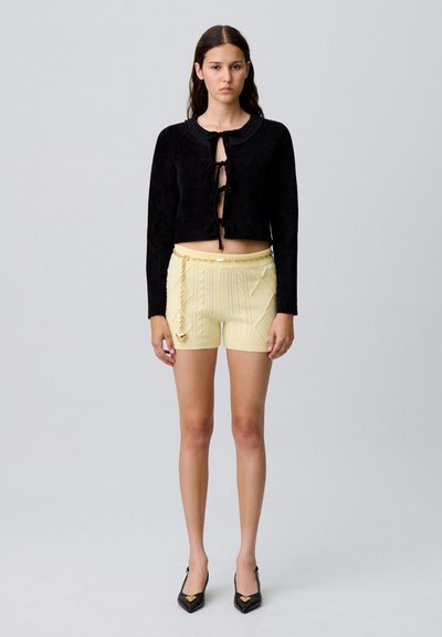 Cárdigan negro corto con cierres de lazo, combinado con pantalones cortos amarillos texturizados con un detalle de cadena. La modelo lleva tacones negros de punta.