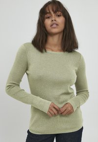 Haut long à manches ribbées de couleur vert clair avec un col rond et une silhouette ajustée. Texture douce, avec des poignets allongés aux poignets.