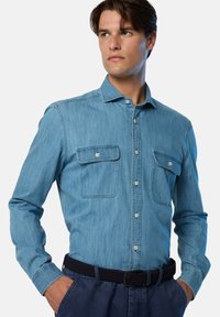 Camicia in denim azzurro chiaro con maniche lunghe, due tasche frontali, colletto con bottoni e chiusura con bottoni. Indossata con una cintura nera intrecciata.
