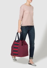 Bolso de mano a rayas rojas y marinas con dos asas, hecho de tela, que presenta un diseño amplio. Llevado por una persona con atuendo casual.