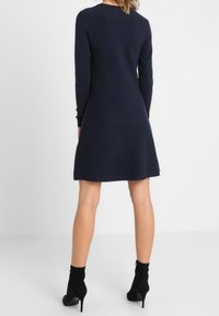 Granatowa sukienka dzianinowa z długim rękawem o rozszerzonym fasonie, okrągłym dekolcie i teksturowanej tkaninie; zestawiona z czarnymi botkami na obcasie.